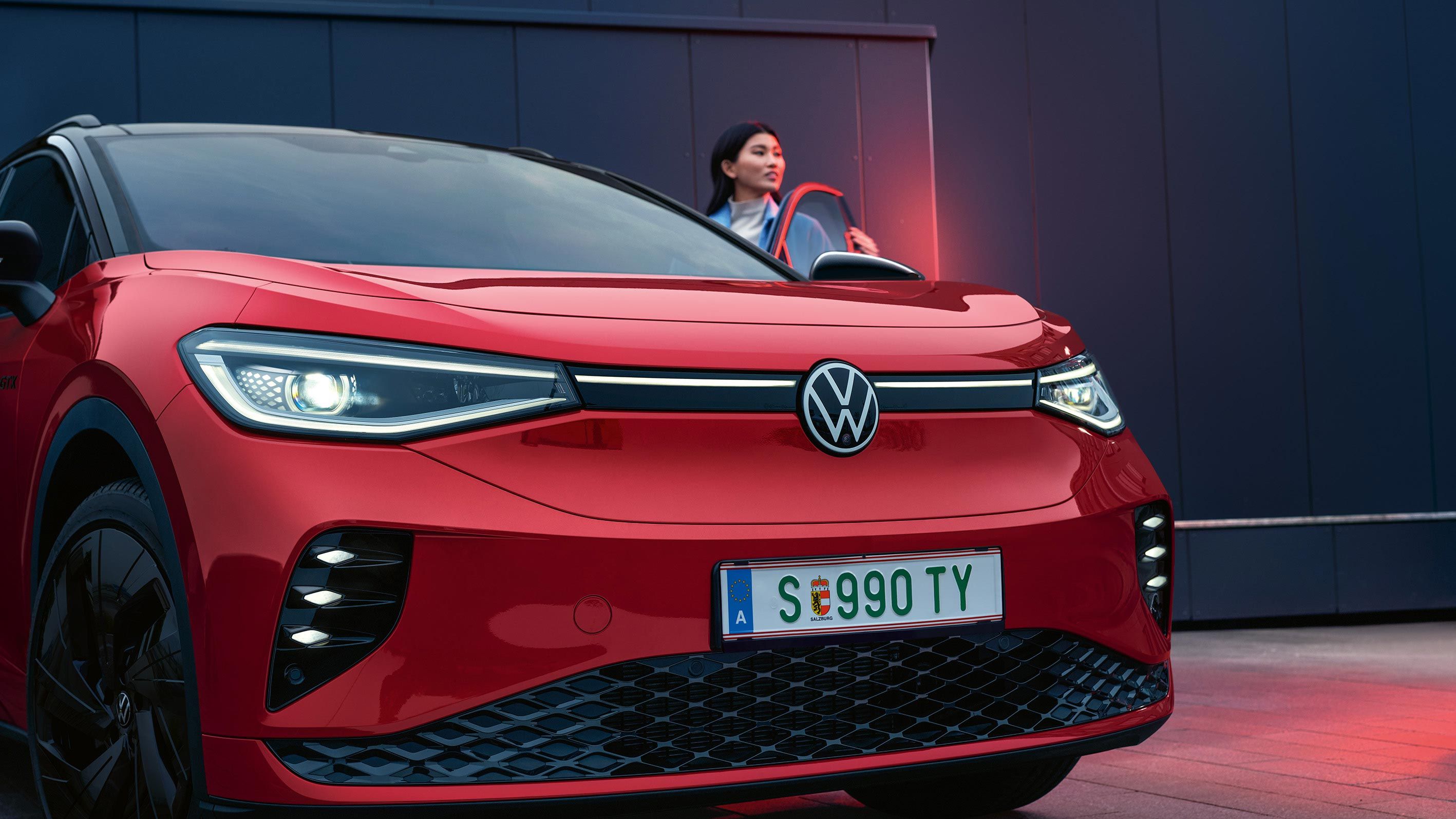 Červený VW ID.4 GTX s otvorenými dverami vodiča. Žena stojí pri dverách a pozerá dopredu.