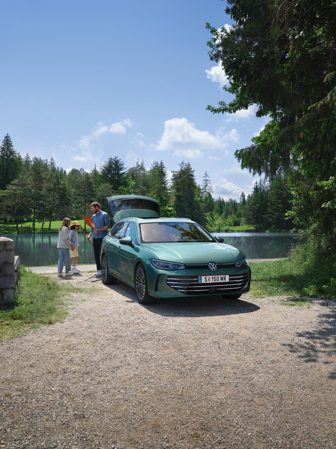 Modrozelený VW Passat s otvoreným kufrom parkuje pri jazere na kúpanie. Muž rozdáva hračky dvom deťom.